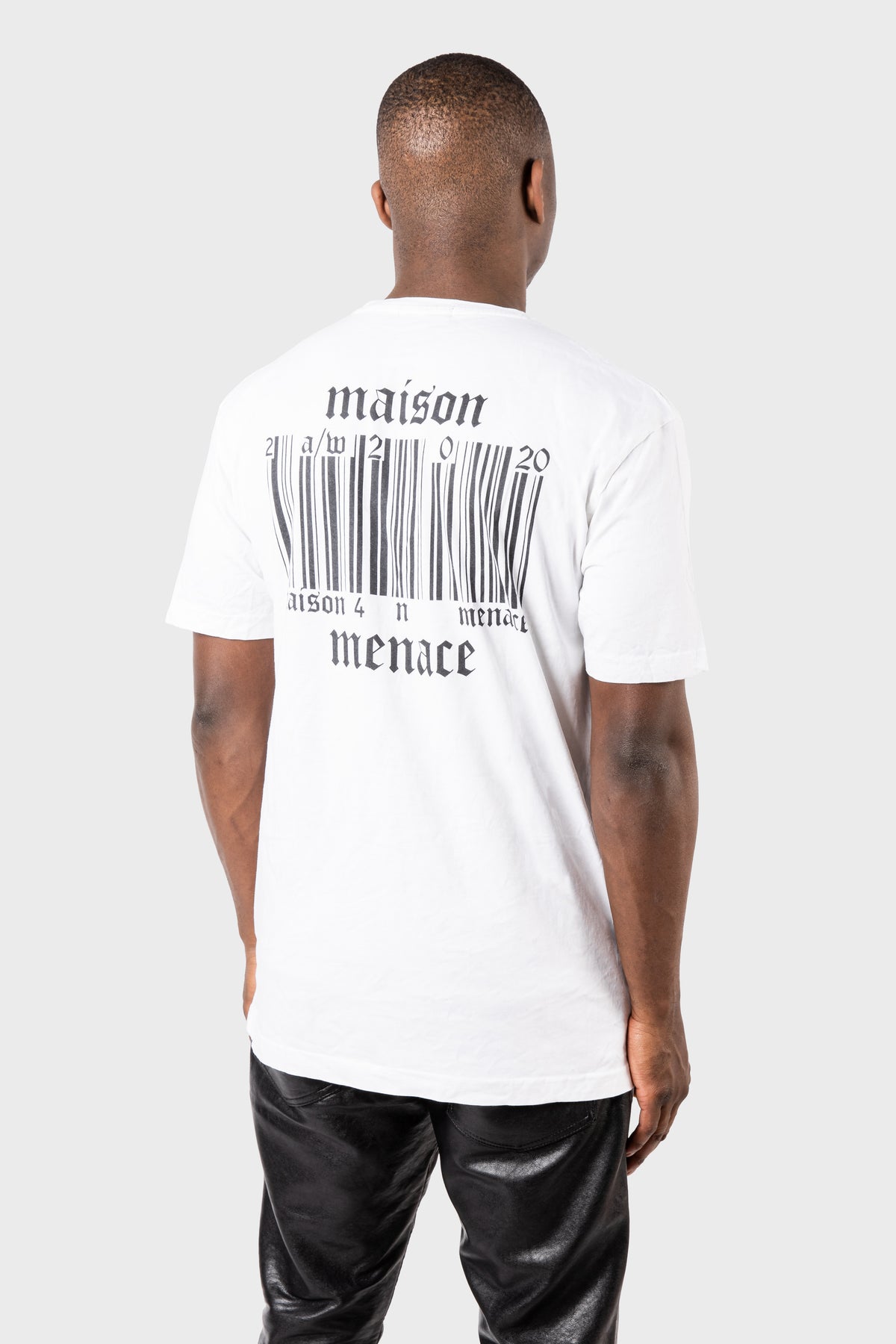 A person with their back to us wears the Broken Barcode T-Shirt from MAISON MENACE, made of organic cotton. The white shirt features a prominent barcode and maison menace text on the back, paired seamlessly with black pants against a plain gray background.