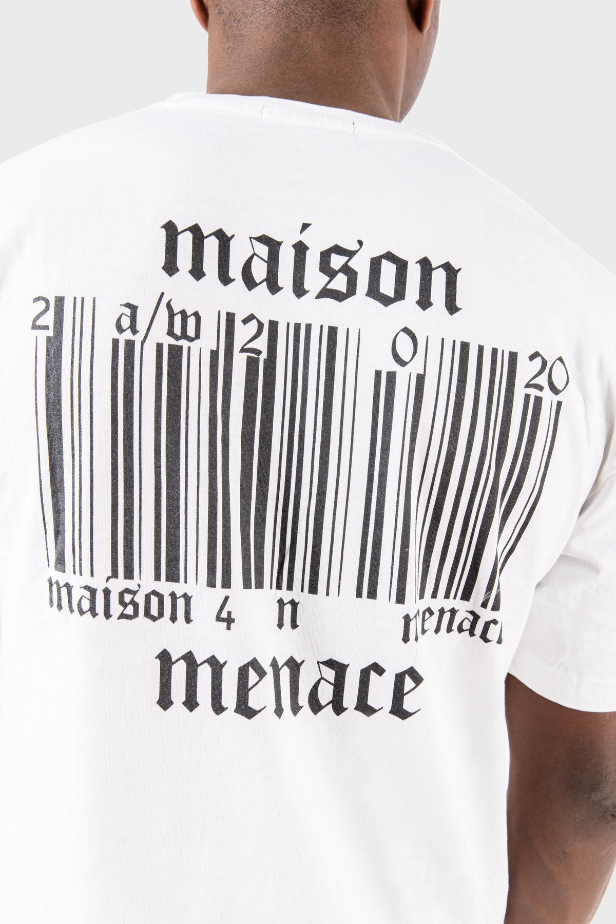A person wearing the Broken Barcode T-Shirt made of soft SUPIMA® cotton by MAISON MENACE, featuring a maison menace logo and distorted barcode graphic on the back, set against a plain gray background for a modern minimalist look.