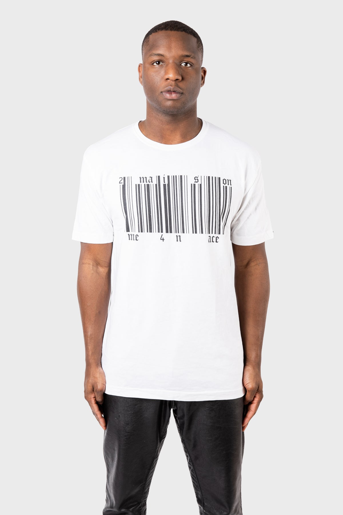 A person in MAISON MENACEs Broken Barcode T-Shirt, made of SUPIMA® cotton with imagination and one 4 all ace text, stands against a plain gray background. They have short hair and are wearing black pants.