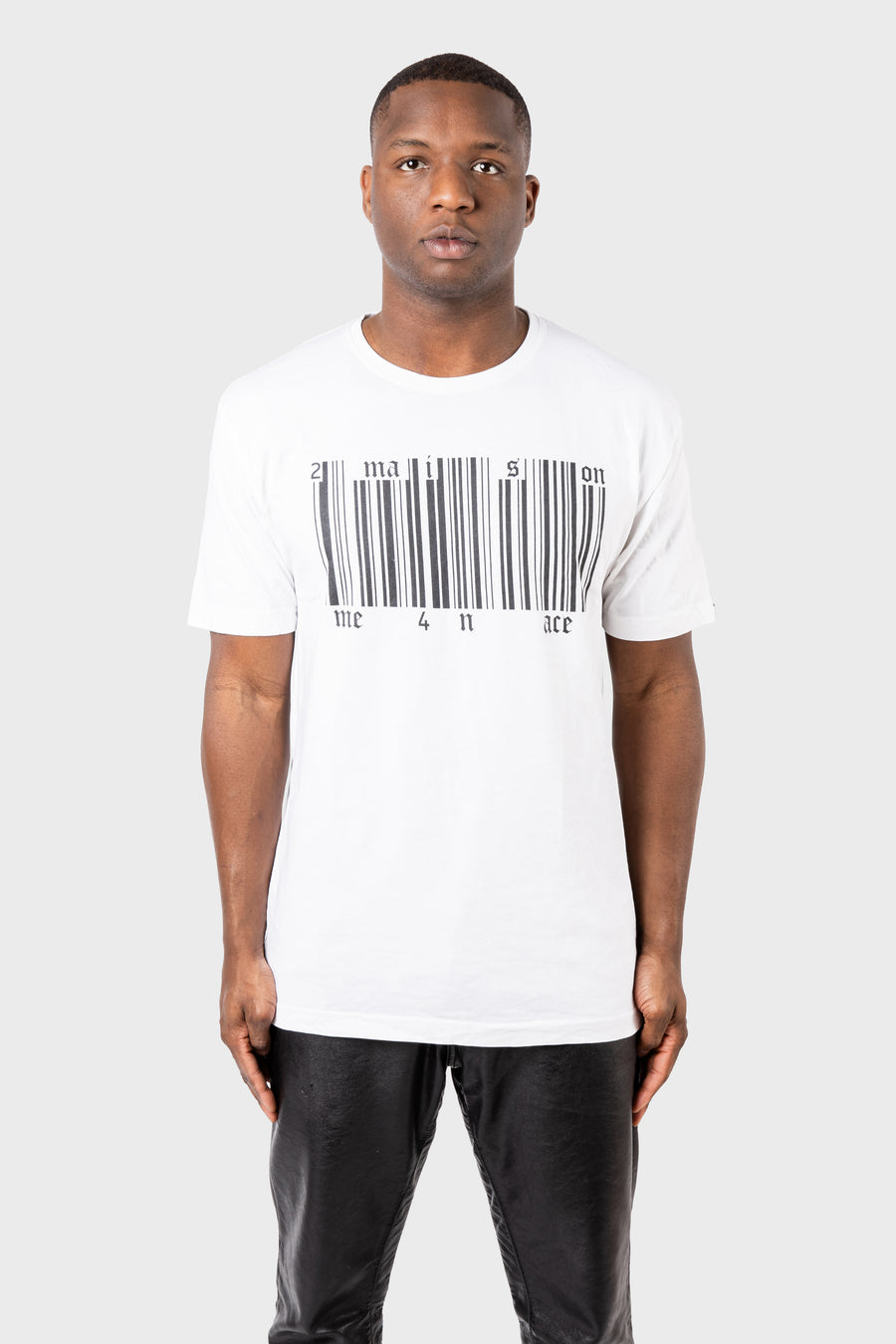 A person in MAISON MENACEs Broken Barcode T-Shirt, made of SUPIMA® cotton with imagination and one 4 all ace text, stands against a plain gray background. They have short hair and are wearing black pants.