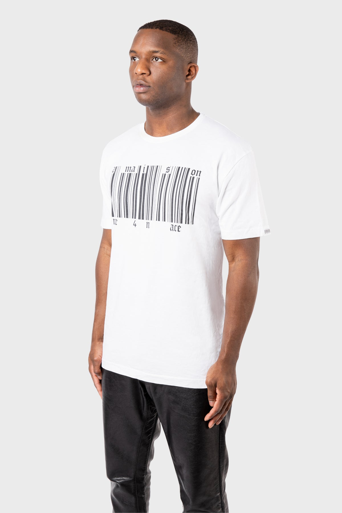 A person wears a MAISON MENACE Broken Barcode T-Shirt in organic cotton with a barcode graphic and black text, paired with black pants, against a plain gray background.