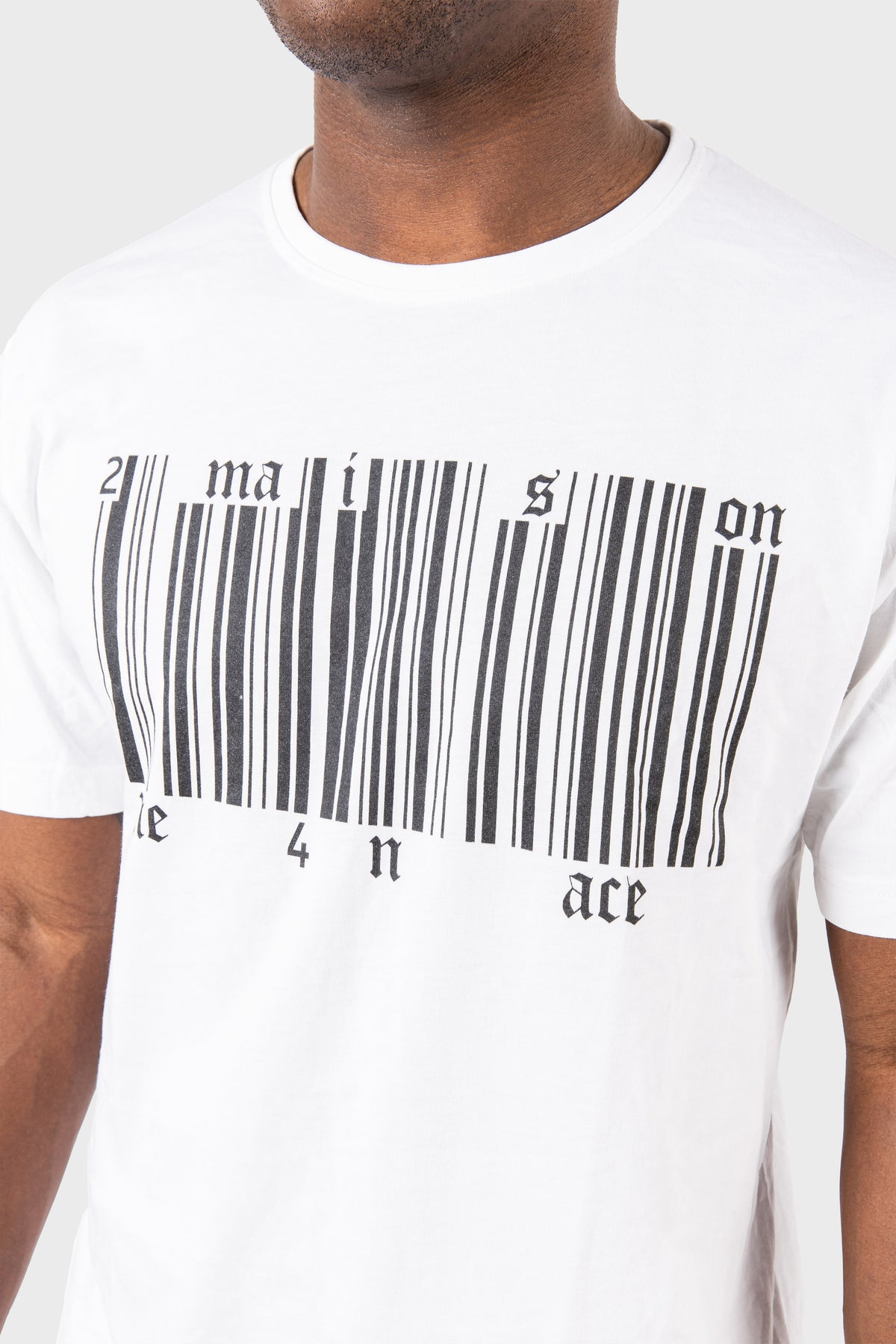 A person wears the MAISON MENACE Broken Barcode T-Shirt, made from luxurious SUPIMA® cotton, featuring a unique barcode graphic that creatively includes maison and ace on a plain gray background.