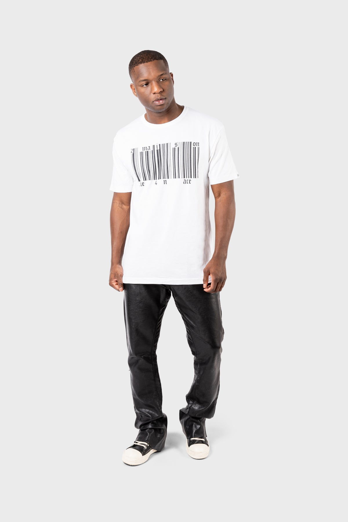 A person wearing a white MAISON MENACE Broken Barcode T-Shirt, black leather pants, and black sneakers stands against a plain gray background, looking slightly to the side.