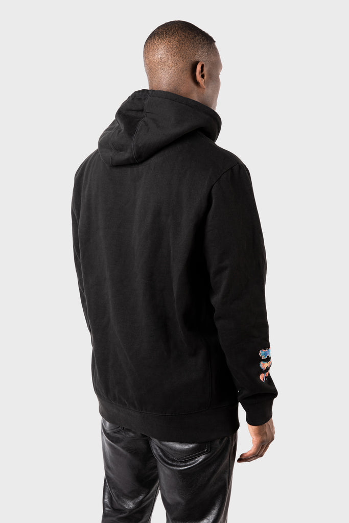A person models a black MAISON MENACE Evolution Hoodie with their back facing the camera against a gray background. The hoodie’s left sleeve showcases small, colorful hearts resembling a watercolor butterfly print, paired with black pants.