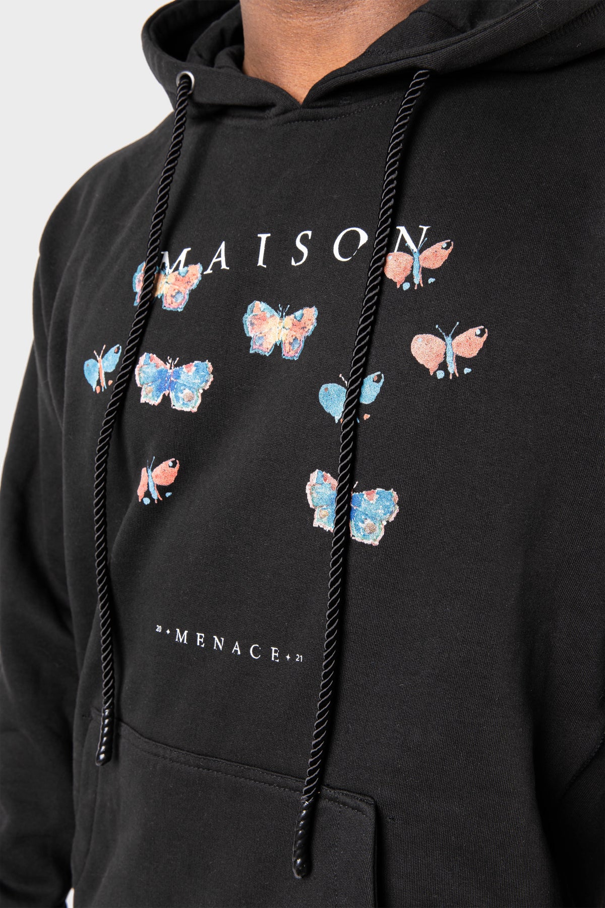 A person wearing the Evolution Hoodie by MAISON MENACE, a heavyweight fleece featuring a watercolor butterfly print, MAISON MENACE text, black drawstrings, and a front pocket.