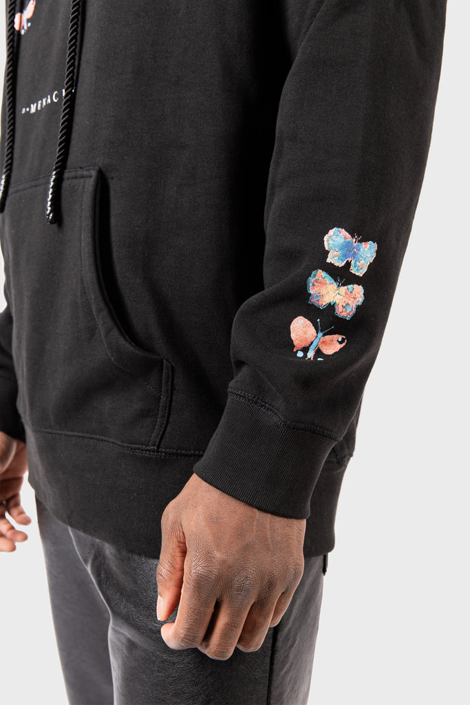 A person wearing the MAISON MENACE Evolution Hoodie in black with a watercolor butterfly sleeve print, front pocket, and drawstrings, paired with dark pants. The background is plain white.