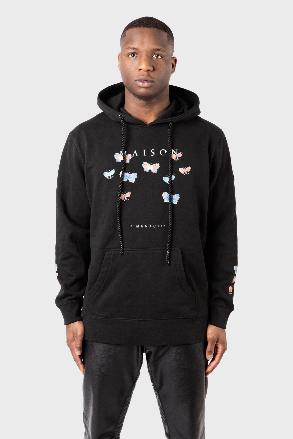 Someone wearing the MAISON MENACE Evolution Hoodie, featuring a vivid watercolor butterfly print and MAISON on the front, stands against a plain background. They pair it with black pants for a striking look.