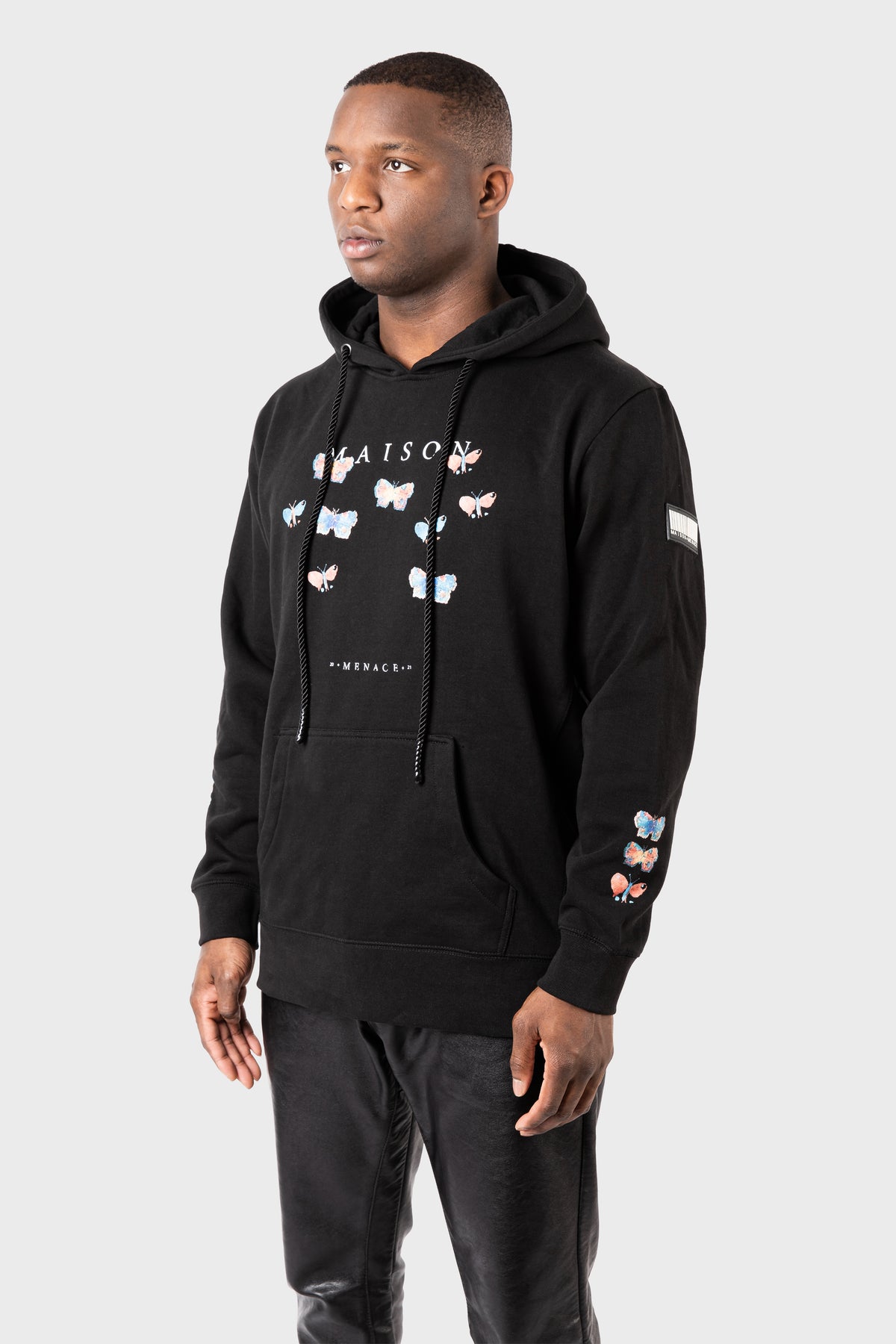 A person stands against a plain gray background wearing the Evolution Hoodie by MAISON MENACE, featuring vibrant watercolor butterfly prints, text on the front, and a patch on the sleeve. They pair it with black pants.