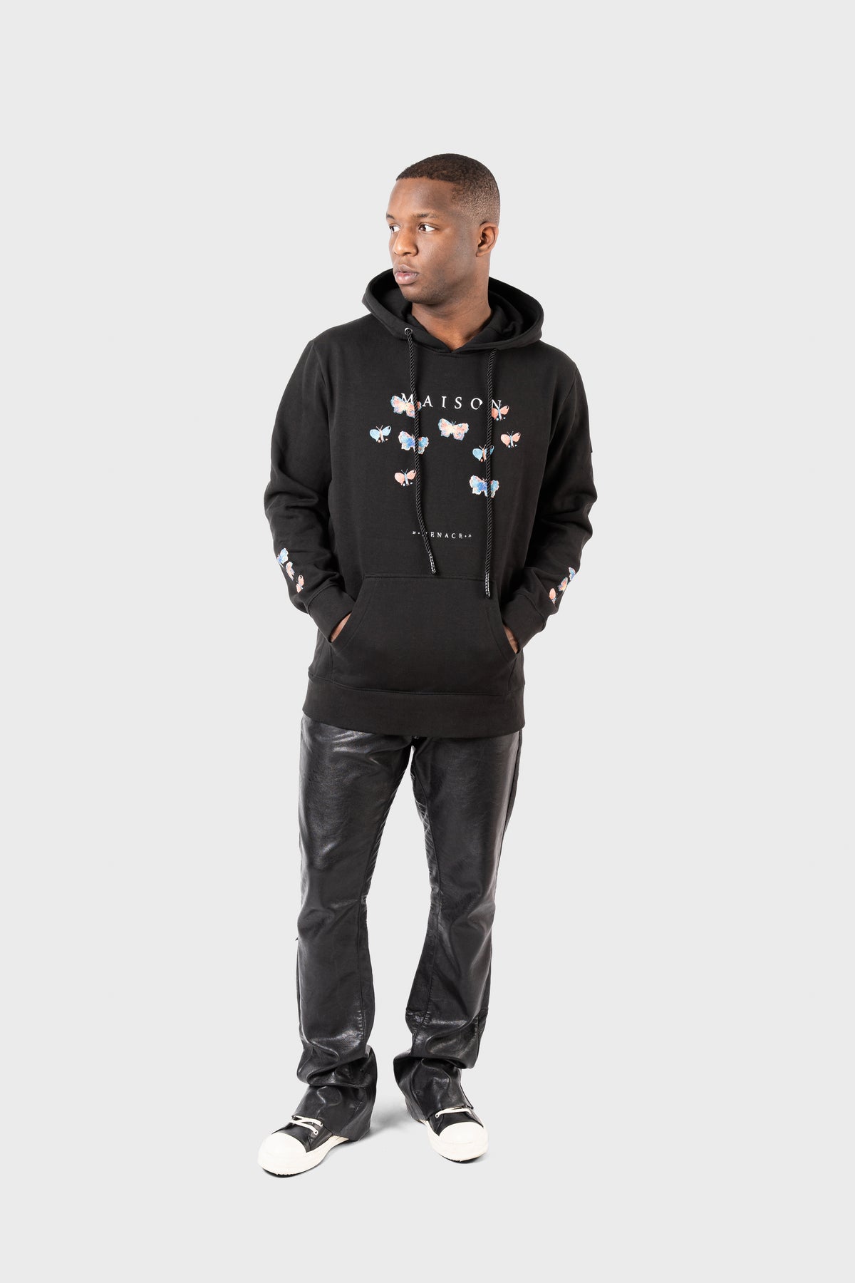 Against a plain backdrop, a person dons the Evolution Hoodie by MAISON MENACE. This black heavyweight fleece hoodie showcases a watercolor butterfly and Maison on the front. Paired with black leather pants and black and white sneakers, they exude effortless style, gazing off to the side.