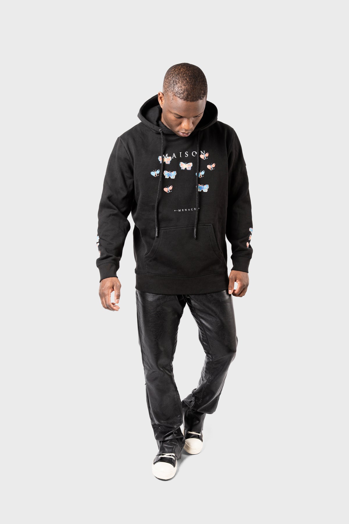 A man wears the MAISON MENACE Evolution Hoodie in black, featuring a watercolor butterfly print and the word MAISON. He pairs it with black pants and black-and-white sneakers, looking down against a plain grey background.