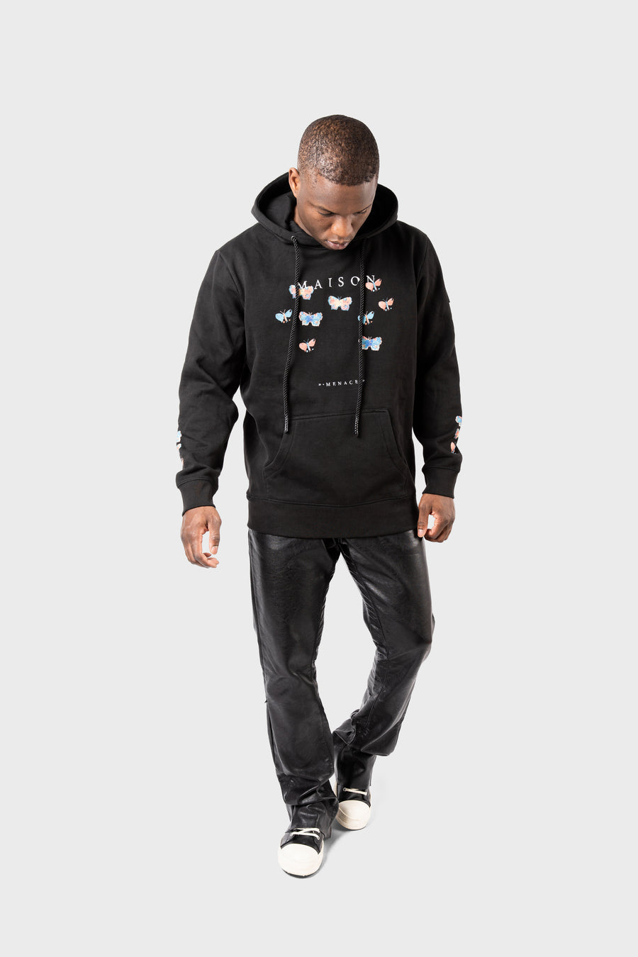 A man wears the MAISON MENACE Evolution Hoodie in black, featuring a watercolor butterfly print and the word MAISON. He pairs it with black pants and black-and-white sneakers, looking down against a plain grey background.