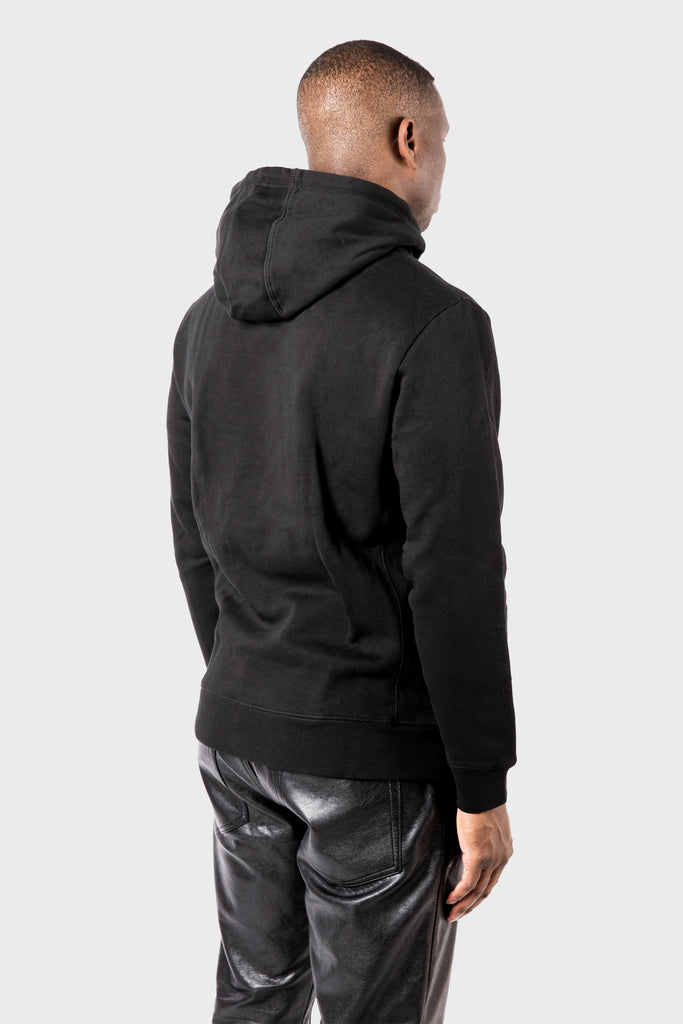 A person stands facing away, wearing a sleek black Noire Classic Hoodie from MAISON MENACE paired with black leather pants, set against a plain gray background that enhances the mysterious allure.
