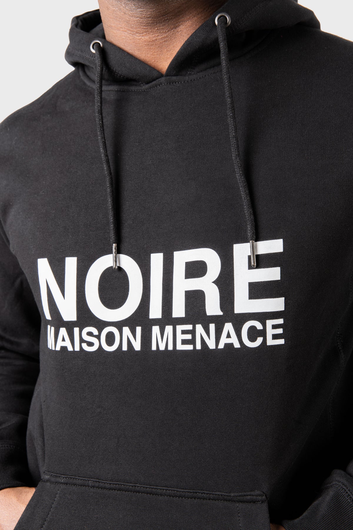 Close-up of a person wearing the Noire Classic Hoodie by MAISON MENACE, featuring bold white letters NOIRE MAISON MENACE on the chest and a drawstring hood, set against a plain white background.