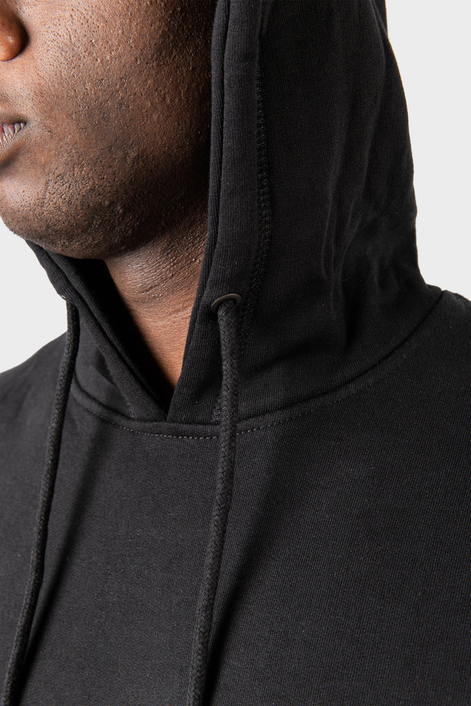 A close-up shows a person wearing the Noire Classic Hoodie by MAISON MENACE with the hood up, focusing on the organic cottons texture and detailed drawstrings. The light background highlights the garments unique quality.