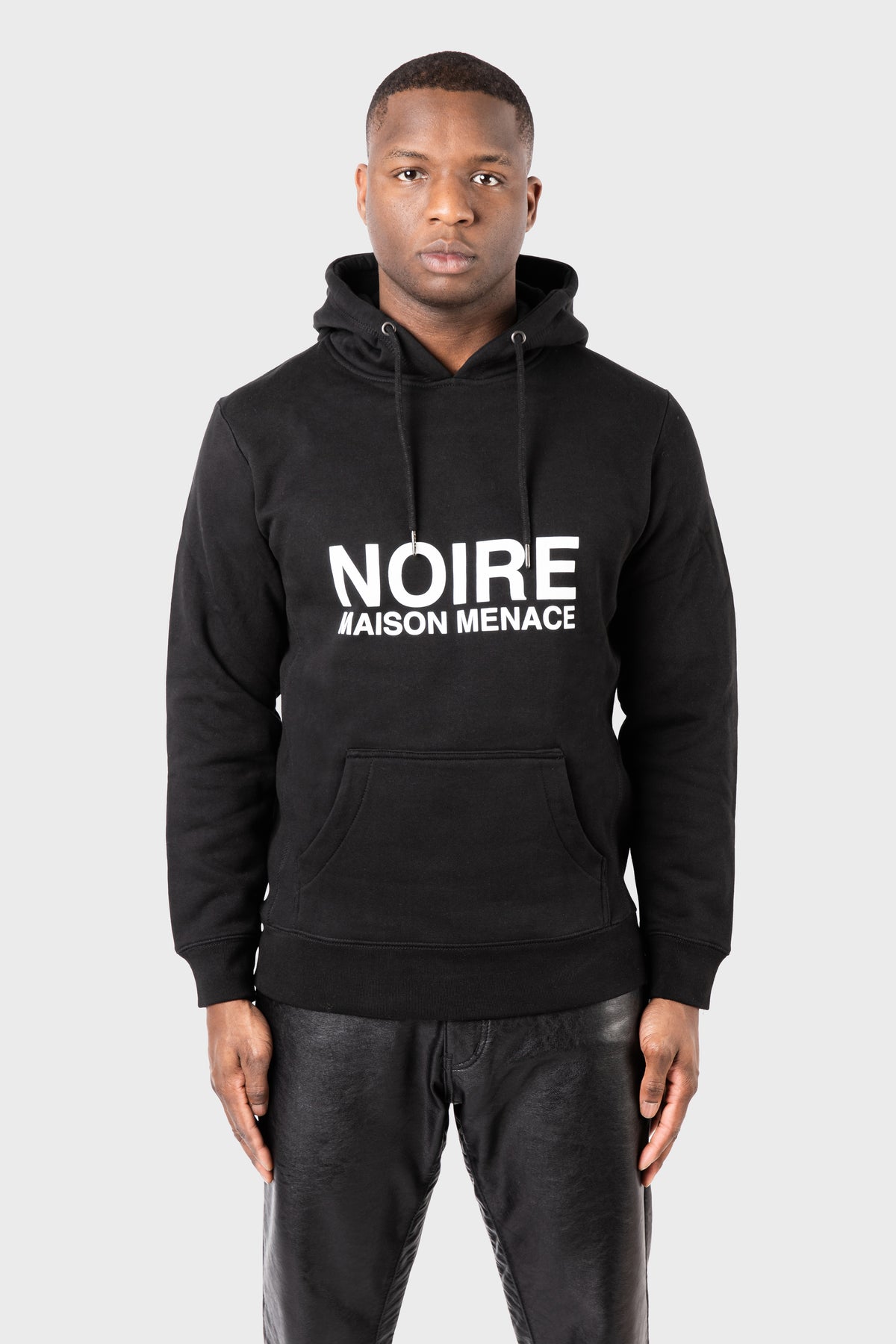 A man in a Noire Classic Hoodie by MAISON MENACE, featuring NOIRE MAISON MENACE prominently on the front, stands against a plain background. Wearing leather pants, he gazes directly at the camera with an understated yet edgy style.