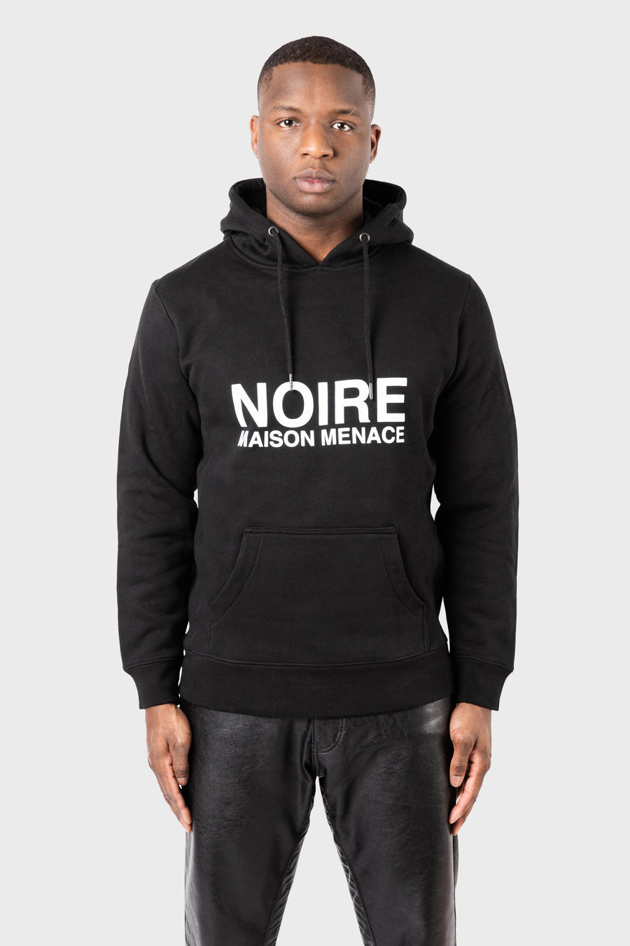 A man in a Noire Classic Hoodie by MAISON MENACE, featuring NOIRE MAISON MENACE prominently on the front, stands against a plain background. Wearing leather pants, he gazes directly at the camera with an understated yet edgy style.