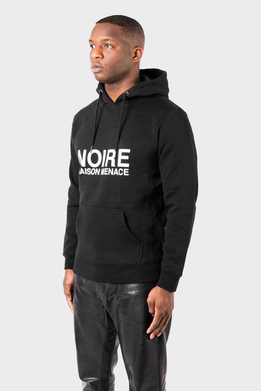 The person is wearing a black Noire Classic Hoodie by MAISON MENACE, featuring NOIRE MAISON MENACE on the front. This organic cotton sweatshirt is paired with black pants against a plain light gray background.