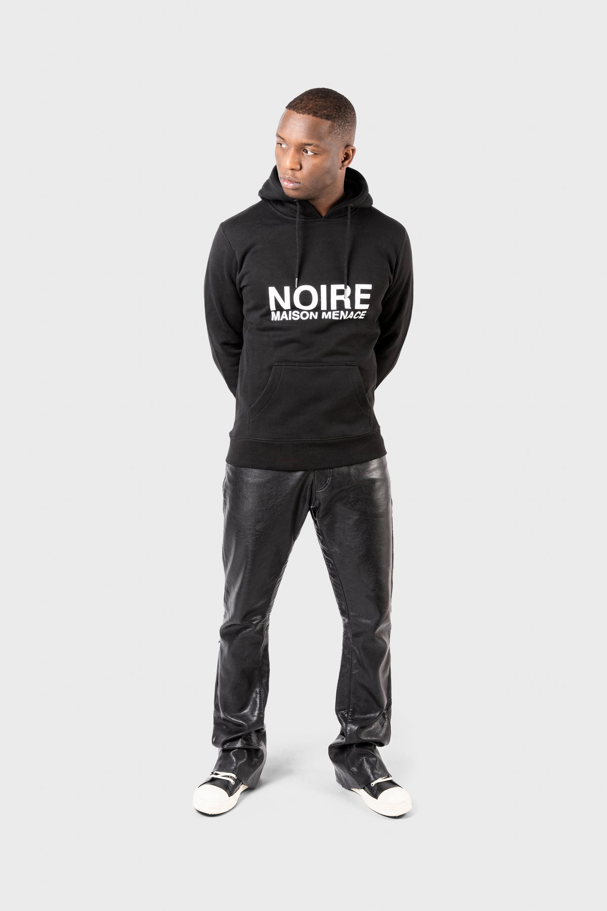 A person stands against a plain background wearing a Noire Classic Hoodie by MAISON MENACE. They look to the side, paired stylishly with black leather pants and monochrome sneakers.