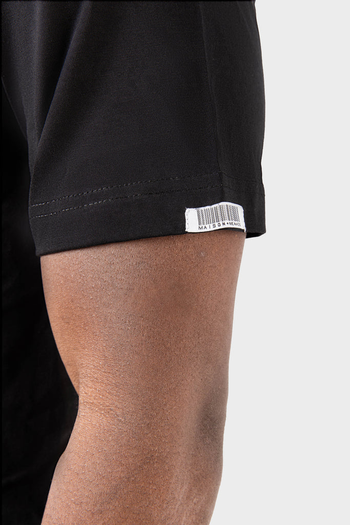 A close-up shows someones arm wearing the Noire Classic T-Shirt by MAISON MENACE, a short-sleeved black shirt made from SUPIMA® cotton. A small white tag with a barcode is near the sleeves edge against a plain gray background.