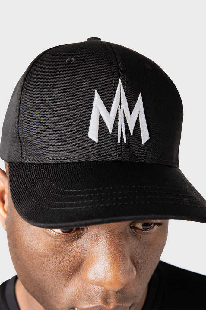 A person wearing a black Maison Menace Signature Hat by MAISON MENACE, crafted from soft cotton twill with a bold white MM logo on the front, stands against a plain light gray background.