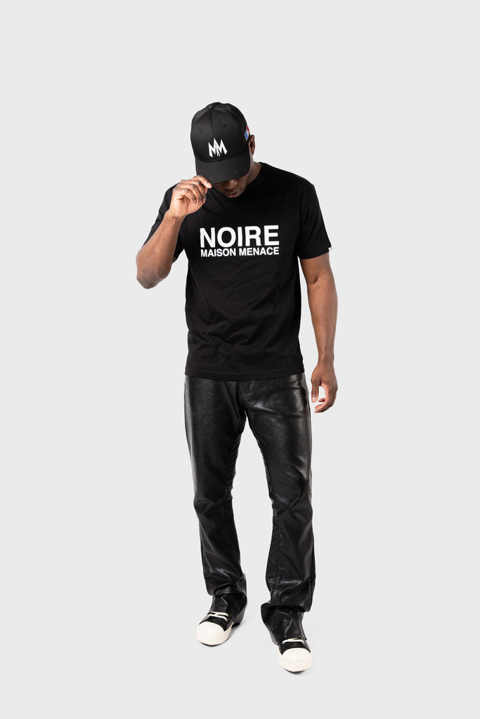 A person wears a Maison Menace Signature Hat in black and a black T-shirt with NOIRE MAISON MENACÉ text, paired with black leather pants and black sneakers featuring white soles, standing against a plain gray backdrop.
