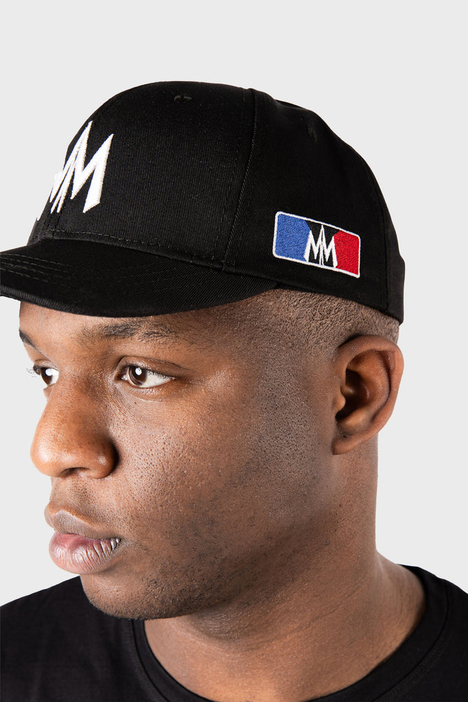 A man wears a Maison Menace Signature Hat in black by MAISON MENACE, made from cotton twill, with MM lettering on the front and a red, white, and blue patch on the side. He looks left with a neutral expression against a plain light gray background.