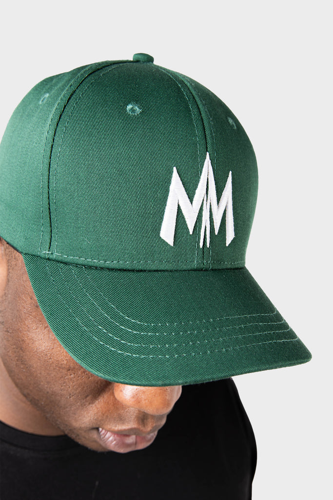 A person in a Maison Menace Signature Hat in Cactus, featuring an embroidered white MM logo, gazes downward against a crisp white background.