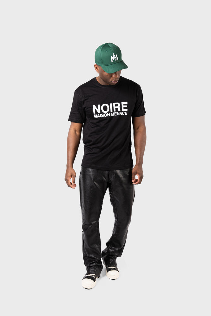 A person wearing a Maison Menace Signature Hat in Cactus with the white MM logo, a black MAISON MENACE T-shirt with NOIRE MAISON MENACE, black leather pants, and black and white sneakers stands against a plain light gray background, looking slightly downward.