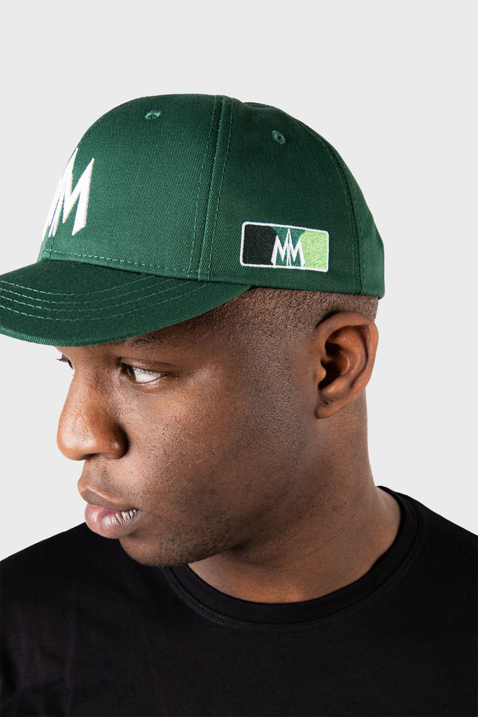 They wear a Maison Menace Signature Hat in Cactus, showcasing a dark green cap with an embroidered white logo and a multicolored rectangular side design. Glancing sideways in a black t-shirt, they stand against a plain light gray background.
