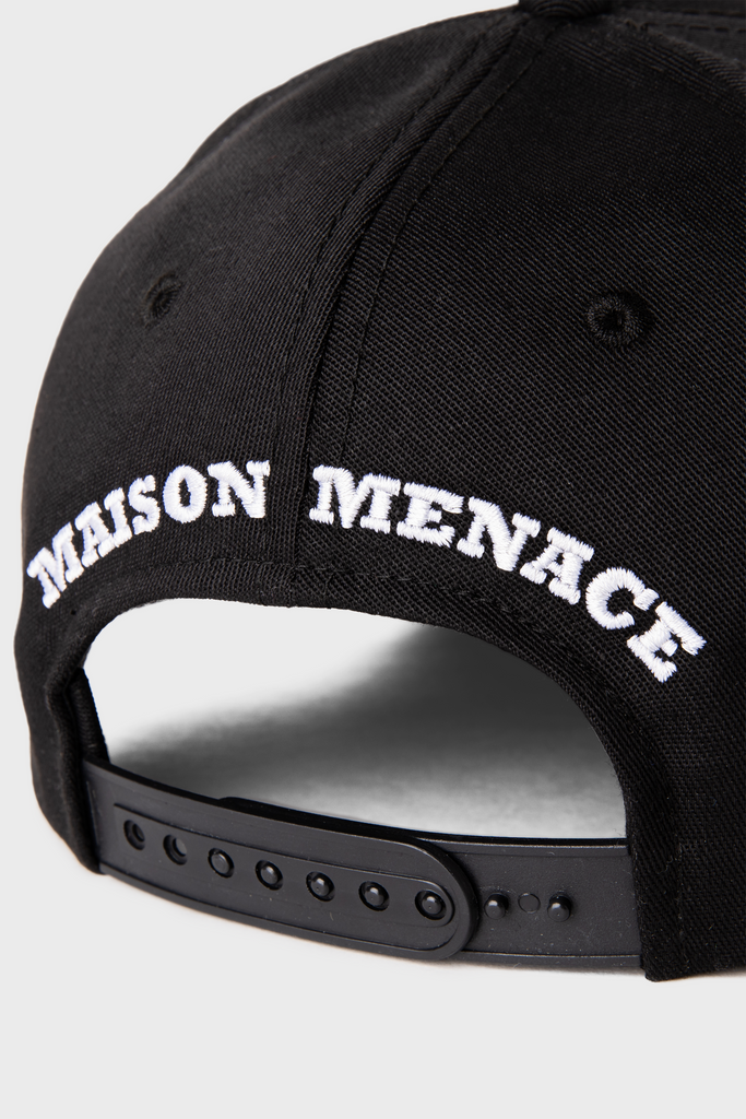 A close-up of the Maison Menace Signature Hat in Black displays its black cotton twill finish, featuring white MAISON MENACE embroidery above an adjustable strap.