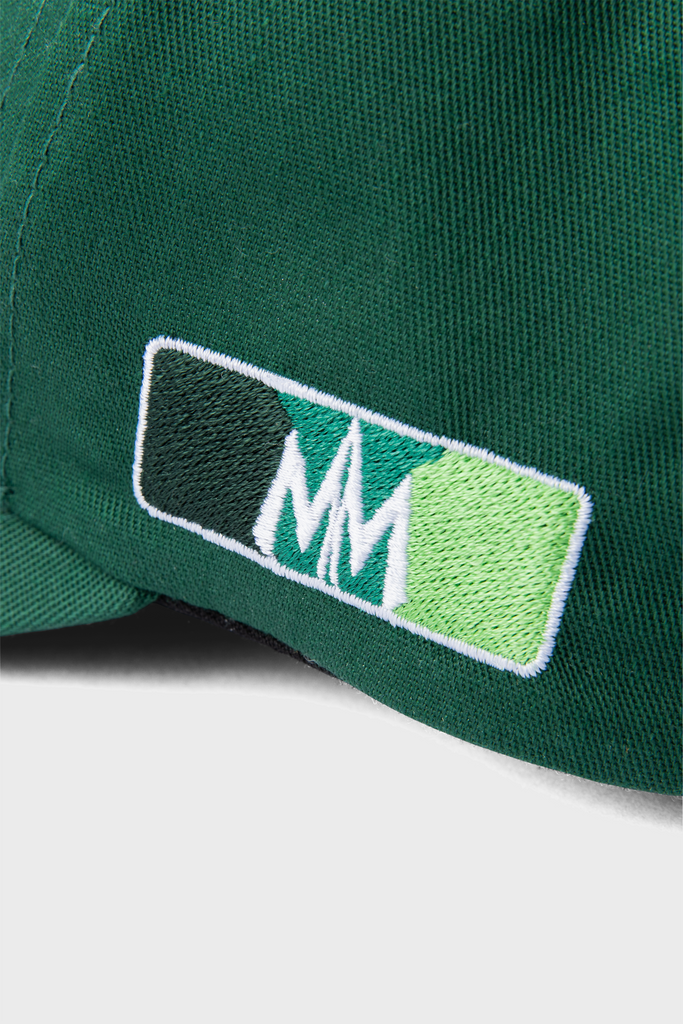 A close-up of a green fabric from MAISON MENACEs Signature Hat in Cactus collection highlights exquisite embroidery with three vertical rectangles in dark green, white, and light green. Overlapping white M letters at the center evoke their signature hat motifs.
