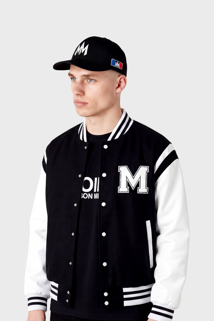 A young man in a black and white varsity jacket with an "M" on the chest, black T-shirt, and the MAISON MENACE Signature Hat in Black stands against a plain light background.