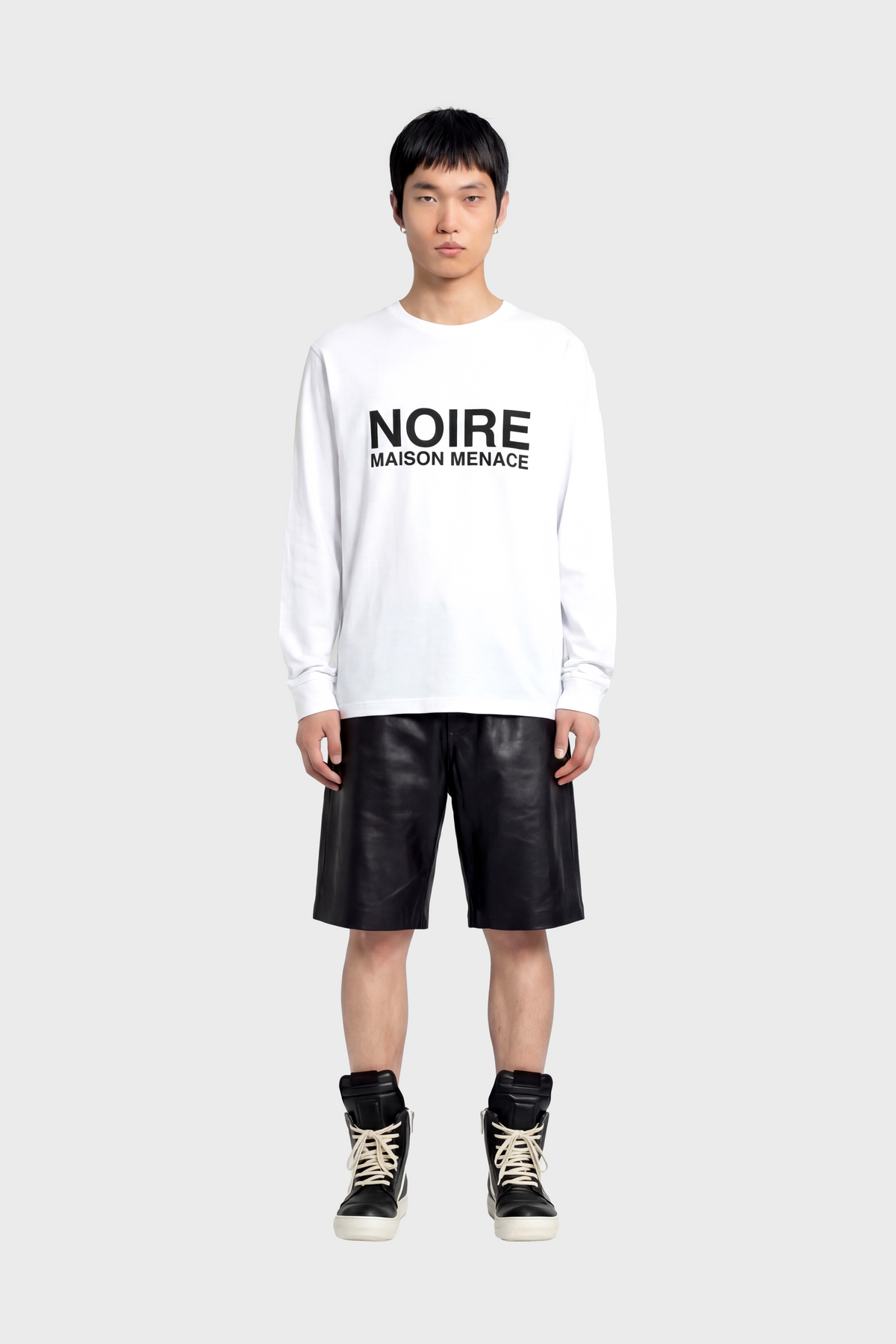 Person wearing a white long sleeve t-shirt with 'NOIRE MAISON MENACE' text, black leather shorts, and high-top sneakers on a white background.