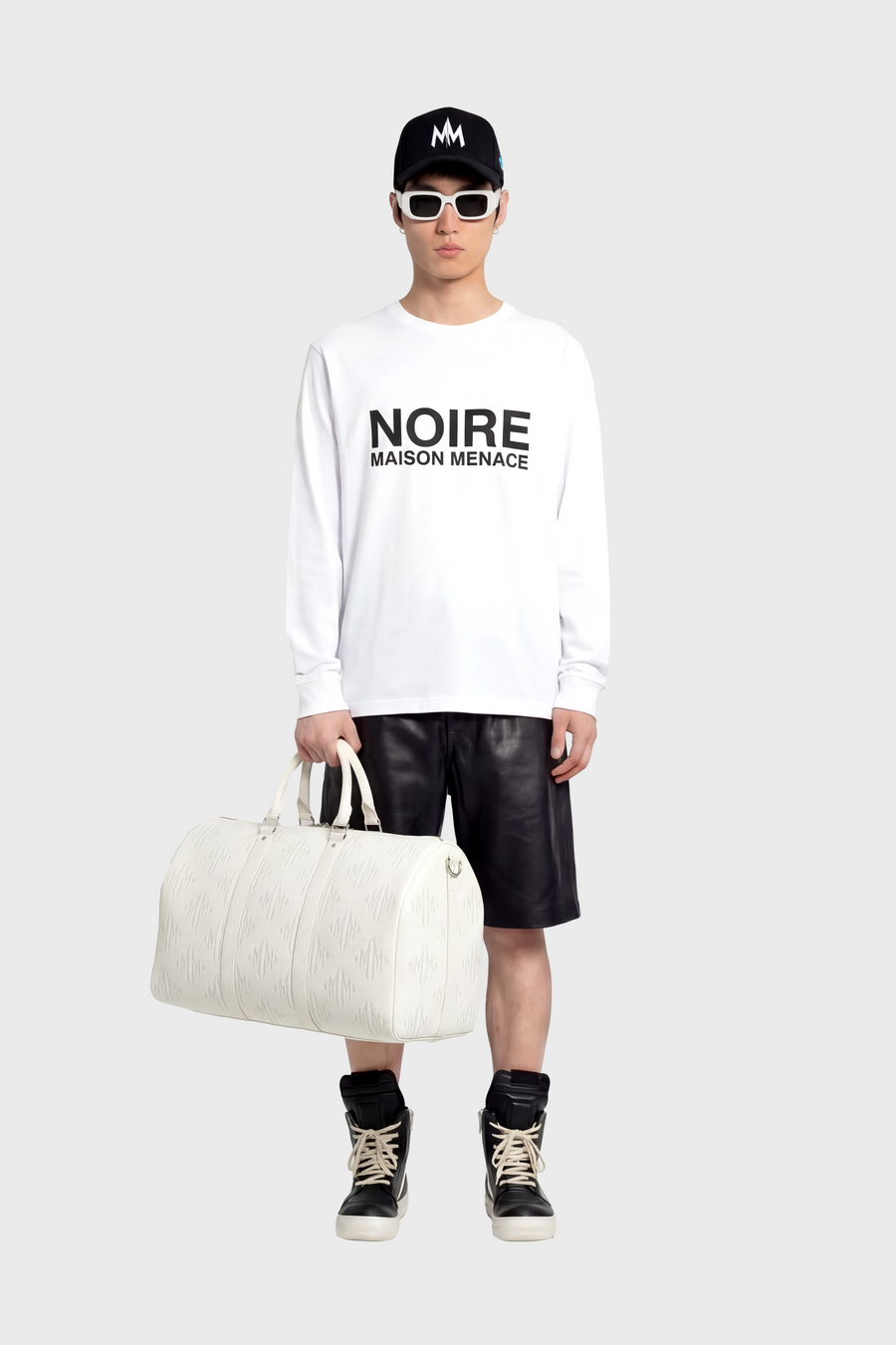 Person wearing a white 'NOIRE' long sleeve t-shirt with black leather shorts and a cap, holding a white bag.