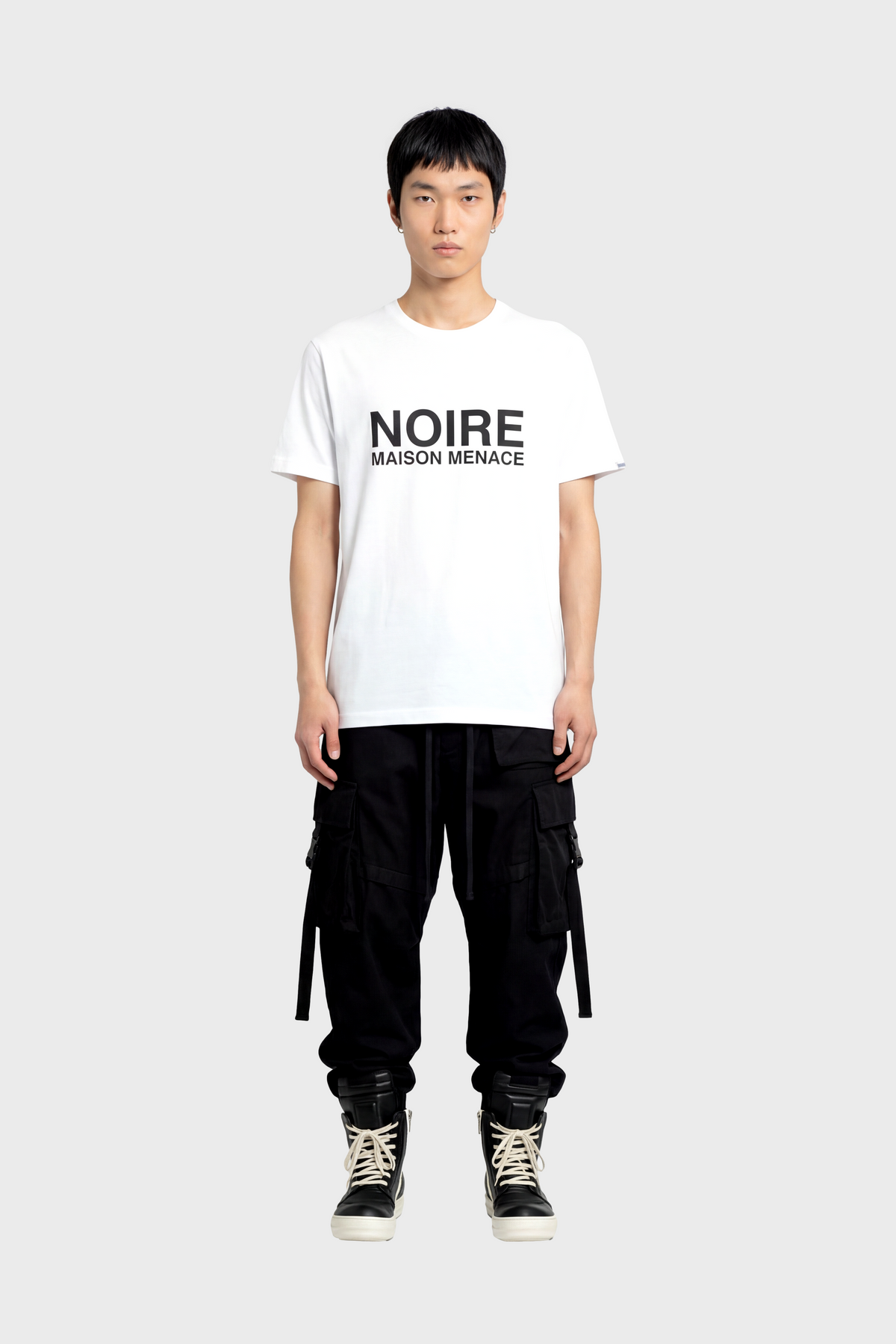 Person wearing a white t-shirt with 'NOIRE MAISON MENACE' text, black cargo pants, and black high-top sneakers on a white background.