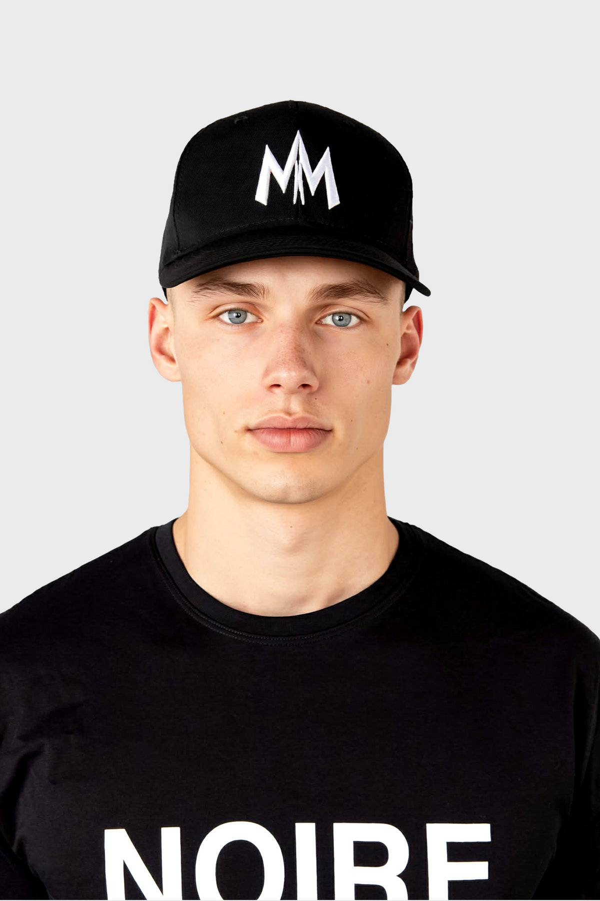 A young man wears the MAISON MENACE Signature Hat in Black and a black T-shirt with NOIRE in white letters, looking directly at the camera against a plain light gray background.