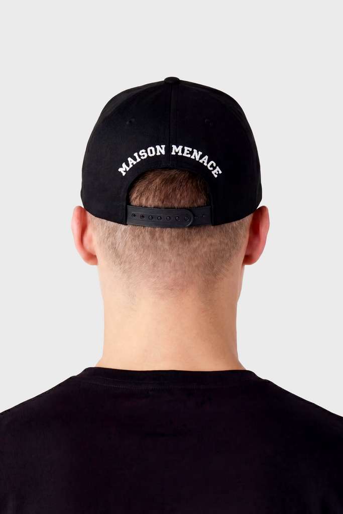 Back view of a person in a black MAISON MENACE Signature Hat, featuring a classic 6-panel design and white MAISON MENACE text on the back, paired with a black shirt against a light grey background.