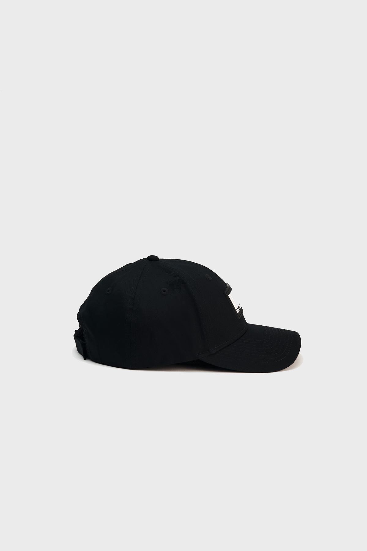 Side view of a black Maison Menace Classic Hat by MAISON MENACE on a light background. This cotton canvas baseball cap has a curved brim, adjustable back strap, and small ventilation holes.