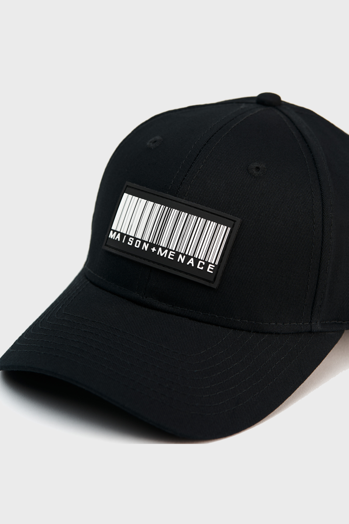 The Maison Menace Classic Hat by MAISON MENACE is a black cotton canvas baseball cap with a barcode patch and text Maison Menace. It features a curved brim, adjustable Velcro closure, and is shown on a neutral backdrop.